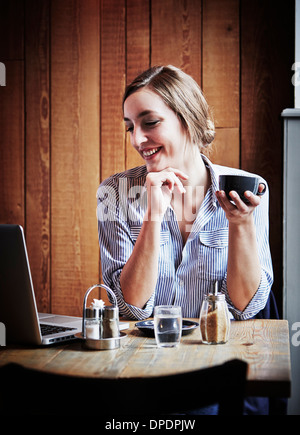 Young woman sitting in cafe holding boisson chaude, à l'aide d'un ordinateur portable Banque D'Images