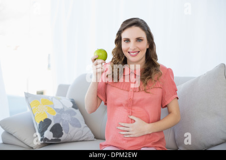 Jeune femme enceinte montrant green apple sitting on couch in living room Banque D'Images