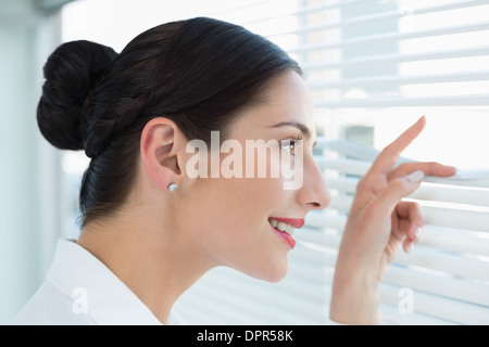 Smiling young business woman peeking through blinds at office Banque D'Images