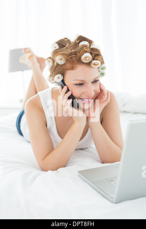 Femme dans les bigoudis using cellphone and laptop in bed Banque D'Images