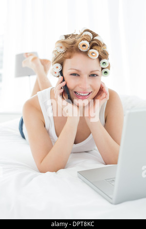 Femme dans les bigoudis using cellphone and laptop in bed Banque D'Images