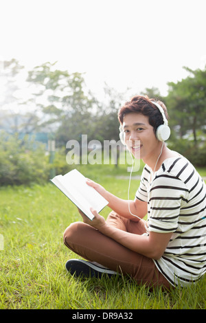 Un homme assis sur l'herbe à écouter de la musique tout en lisant un livre Banque D'Images