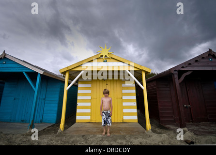 Un jeune garçon se tient à l'extérieur d'une cabane de plage de couleur vive dans la grande l'été britannique Banque D'Images