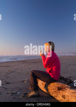 Une femme blonde, apprécie son verre de vin blanc, assis sur un journal de dérive, à la plage, Hokitika, New Zealand. Banque D'Images