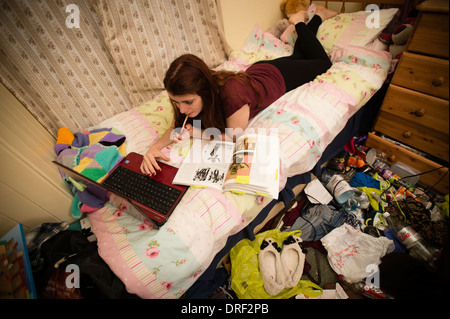 Une jeune femme university student lying on her bed étudier travailler lecture écriture sur ordinateur portable dans la chambre en désordre désordre à la maison UK Banque D'Images