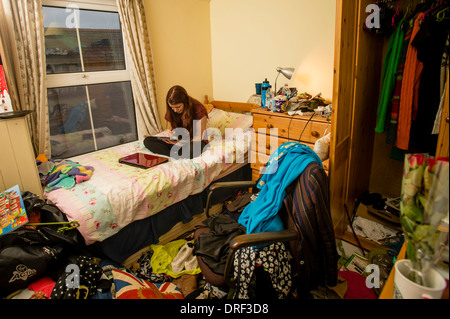 Une jeune femme étudiant de l'université travaillant sur son iPad dans sa chambre en désordre à la maison UK Banque D'Images