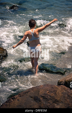 Femme d'âge moyen de marcher dans l'eau sur la plage de rochers. Banque D'Images