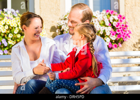 Famille avec la mère, le père et la fille ensemble sur banc de jardin en face de la maison Banque D'Images