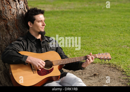 Un jeune homme assis contre un arbre dans un parc à jouer de la guitare Banque D'Images