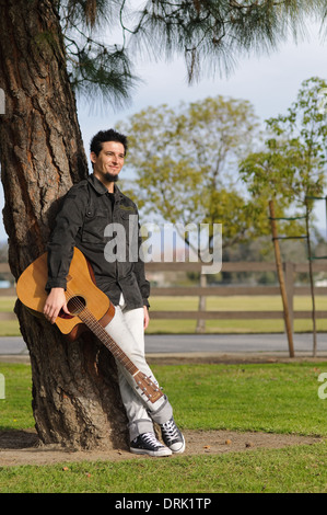 Jeune homme, musicien debout par un arbre dans un parc tenant une guitare et smiling Banque D'Images