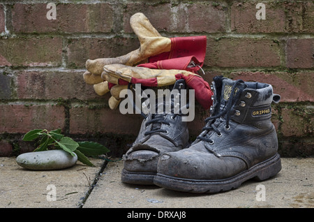Une paire de vieilles bottes de travail dans le jardin par un mur avec une paire de gants de jardinage Banque D'Images