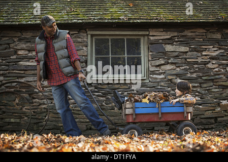 Un homme en veston rembourré tirant un panier de jardin avec un enfant assis parmi l'automne les feuilles séchées d'un bâtiment traditionnel en pierre Banque D'Images