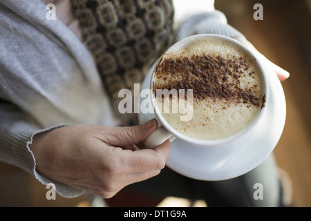 Une personne titulaire d'une pleine tasse de café cappuccino mousseux dans un livre blanc la chine tasse chocolat en poudre saupoudré dans un motif Banque D'Images