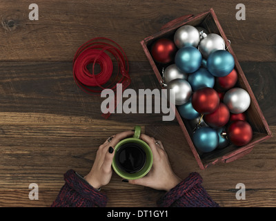 Bleu Rouge et d'argent des ornements et ruban rouge dans une boîte sur une planche de bois une main enroulée autour d'une tasse de café en faisant une pause Banque D'Images