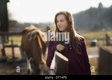 Une fille s'appuyant sur une clôture de pâturage et un cheval dans l'arrière-plan sur une ferme biologique Banque D'Images