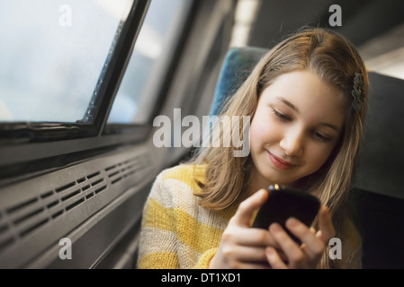 Une jeune fille aux longs cheveux blonds assis par une fenêtre d'un train à l'aide de son téléphone mobile et l'envoi de messages sms Banque D'Images