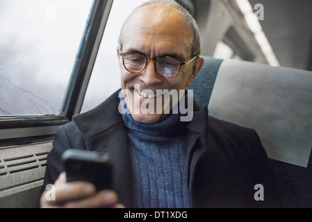 Un homme mûr assis par une fenêtre dans un train à l'aide de son téléphone portable de rester en contact en déplacement Banque D'Images