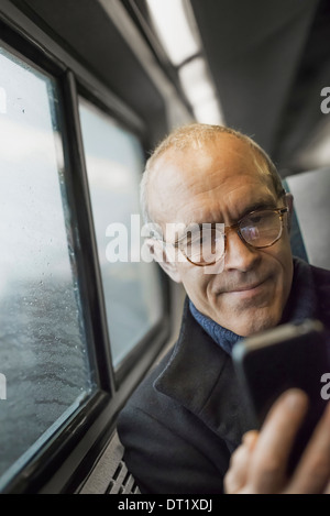 Un homme mûr assis par une fenêtre dans un train à l'aide de son téléphone portable de rester en contact en déplacement Banque D'Images