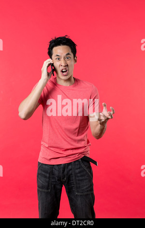 Angry Asian man talking on cell phone. L'article contre l'arrière-plan rouge. Banque D'Images