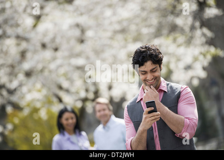 Les gens à l'extérieur dans la ville au printemps fleur de cerisier sur les arbres un homme contrôler son téléphone cellulaire et deux personnes derrière lui Banque D'Images