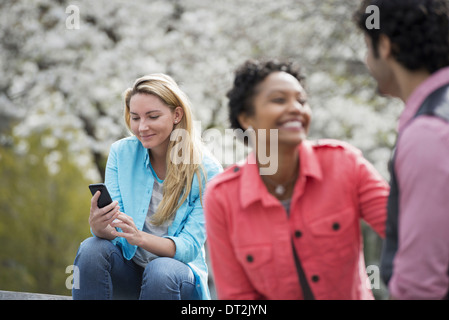 Spring Time New York City park White blossom sur les arbres d'une femme assis sur un banc tenant son téléphone mobile un couple à côté d'elle Banque D'Images
