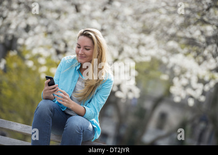 Spring Time New York City park White blossom sur les arbres une femme assise sur un banc tenant son téléphone mobile Banque D'Images