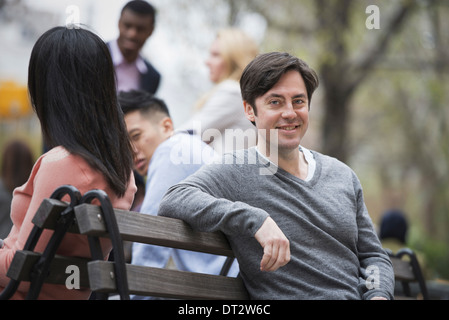 Vue sur citySitting sur un banc de parc un homme souriant à l'appareil photo quatre personnes dans l'arrière-plan Banque D'Images
