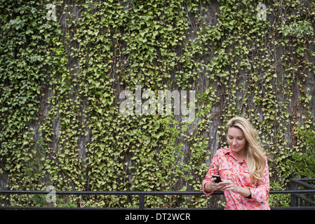 Parc de la ville de printemps avec un mur recouvert de plantes grimpantes et de lierre une jeune femme aux cheveux blonds contrôler son smart phone Banque D'Images