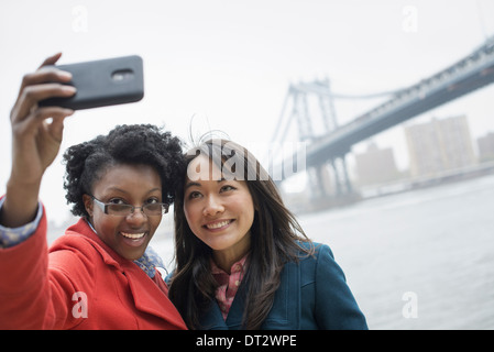 Le Pont de Brooklyn, traversée de la rivière de l'est un couple de deux femmes de prendre une photo avec un téléphone un selfy d'eux-mêmes Banque D'Images