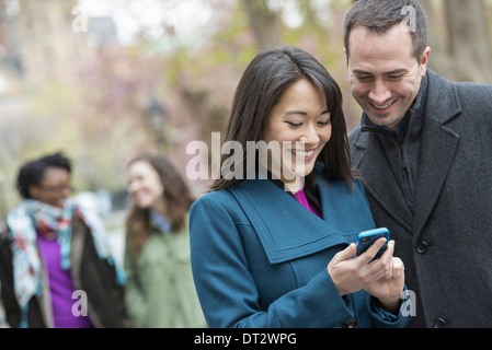 Un groupe de personnes dans un parc de la ville un homme dans un manteau gris et une femme dans un manteau turquoise à la fois à la recherche d'un téléphone intelligent Banque D'Images