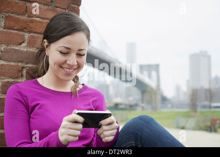 Passage sur le pont de Brooklyn, l'East River, une femme assise appuyée contre un mur de briques contrôler son smart phone Banque D'Images