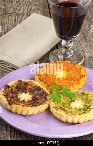 Assortiment de mini quiches végétariennes, servis sur une plaque rose et un verre de vin rouge sur un fond de bois Banque D'Images