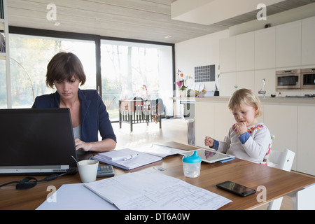 Mère avec enfant de sexe féminin travaillant sur ordinateur à la table de cuisine Banque D'Images