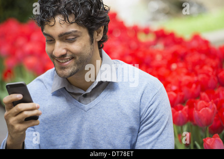 Mode de vie urbain. Un homme dans le parc, à l'aide de son téléphone mobile. Un lit de tulipes fleurs rouges en arrière-plan. Banque D'Images