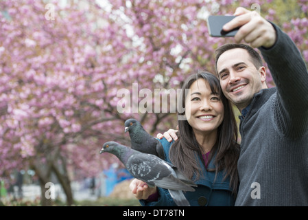 Un couple, un homme et une femme, dans le parc, en prenant un selfy, self portrait avec un téléphone mobile. Deux pigeons perché sur son poignet. Banque D'Images