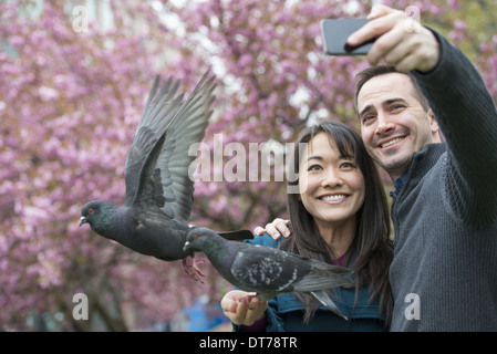 Un couple, un homme et une femme, dans le parc, en prenant un selfy, self portrait avec un téléphone mobile. Deux pigeons perché sur son poignet.
