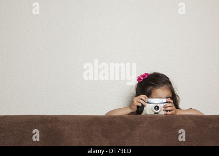 Une jeune fillette de trois ans accroupi derrière un canapé, avec un appareil photo jouet, de prendre une photo. Banque D'Images