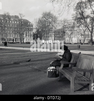 Années 1950, tableau historique d'une dame assise seule sur un banc de parc en bois à Hyde Park, Londres, Angleterre la lecture d'un livre. Banque D'Images