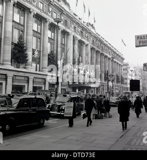 Années 1960, historique, décorations de Noël sur le grand magasin Selfridges sur une rue animée d'Oxford Street, Londres, Angleterre. Circulation très fréquentée avec taxis londoniens dans la rue. Sur le trottoir, un homme avec une planche sandwich. Banque D'Images