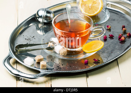 Tasse de thé en verre avec des morceaux de sucre, de citron et de boutons de rose, servi sur plateau vintage argent sur table en bois blanc. Banque D'Images