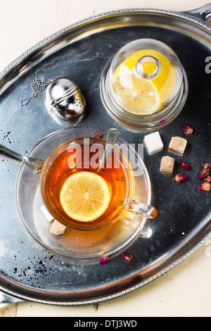 Vue de dessus sur verre tasse de thé avec des morceaux de sucre, de citron et de boutons de rose, servi sur plateau vintage argent sur table en bois blanc. Banque D'Images