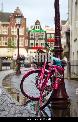 Un vélo rose appuyé contre une lampe de rue après la pluie. Le sol est humide. Il y a des maisons typiquement néerlandais à l'arrière-plan. Banque D'Images