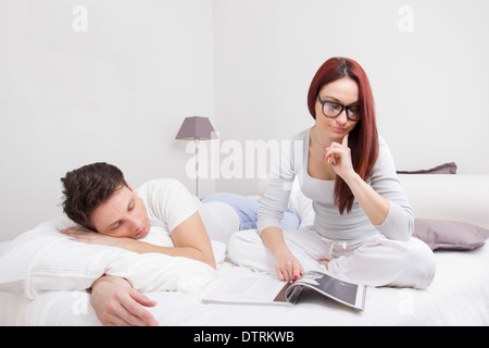 Couple au lit où le jeune femme avec des lunettes est en train de lire un livre et l'homme est en train de dormir à côté d'elle Banque D'Images