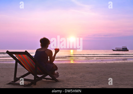 Femme d'âge moyen avec cocktail admirer le coucher du soleil sur la plage Banque D'Images