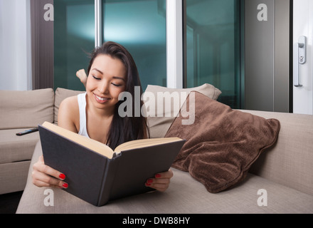 Happy young woman reading book while lying on sofa Banque D'Images