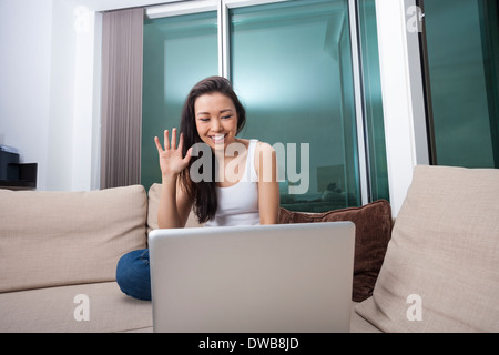 Smiling young woman waving en face de l'ordinateur portable sur canapé Banque D'Images