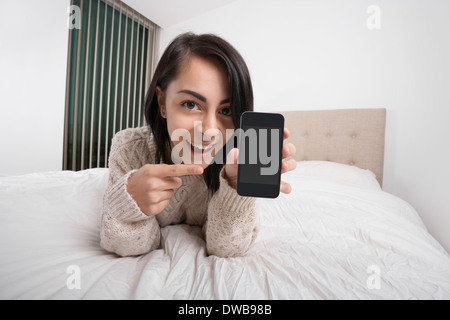 Portrait of happy woman pointing at smart phone while Lying in Bed Banque D'Images