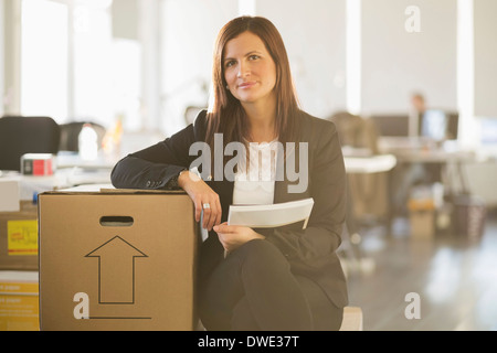 Portrait of young businesswoman sitting on cardboard box in new office Banque D'Images