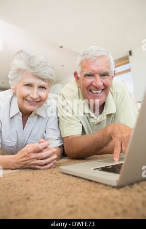Cheerful senior couple en utilisant l'ordinateur portable ainsi allongé sur un tapis Banque D'Images