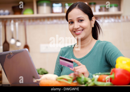 Smiling woman magasinage en ligne à l'aide d'ordinateur et une carte de crédit dans la cuisine Banque D'Images